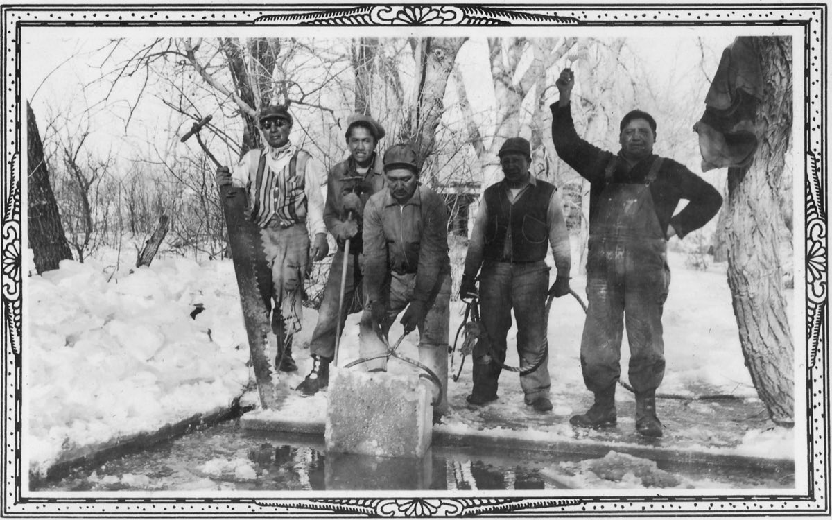 Five men cutting large blocks of ice from a frozen lake with saws and hooks.