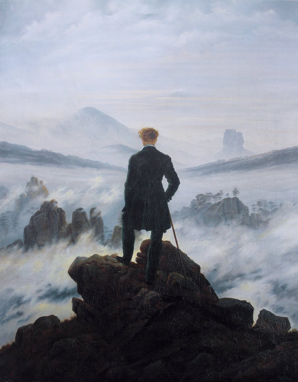 A man seen from behind stands on a dark rocky summit, looking out over a sea of fog and faint distant peaks.