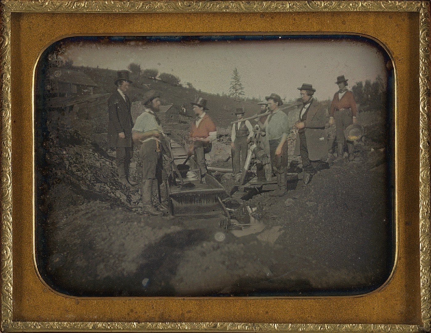 A group of California Gold Rush miners posed at a wooden sluice called a long tom, shovels and pans in hand, beside a diverted stream.