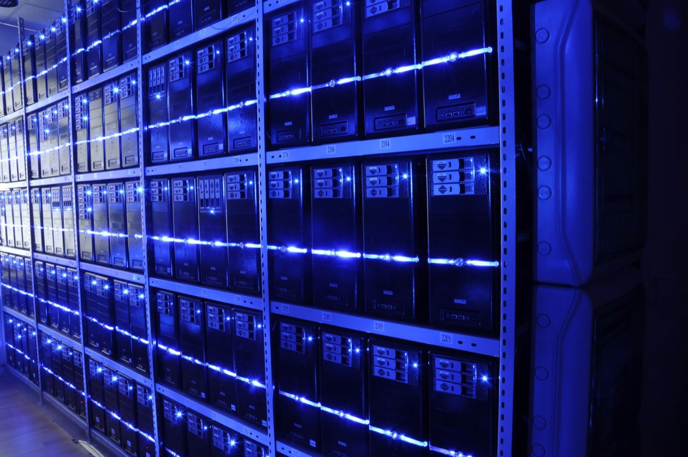 A long aisle inside a modern data center, rows of identical server racks receding under cool overhead light with bundled cabling overhead.