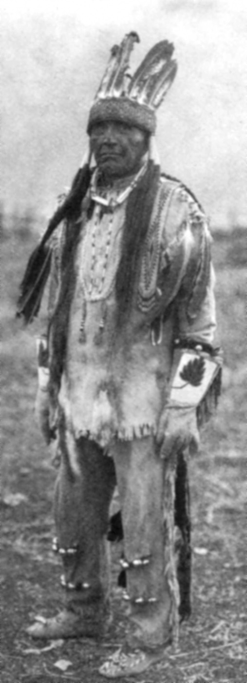 A full-length portrait of a Klamath man in traditional dress photographed by Edward S. Curtis, standing and looking toward the camera.