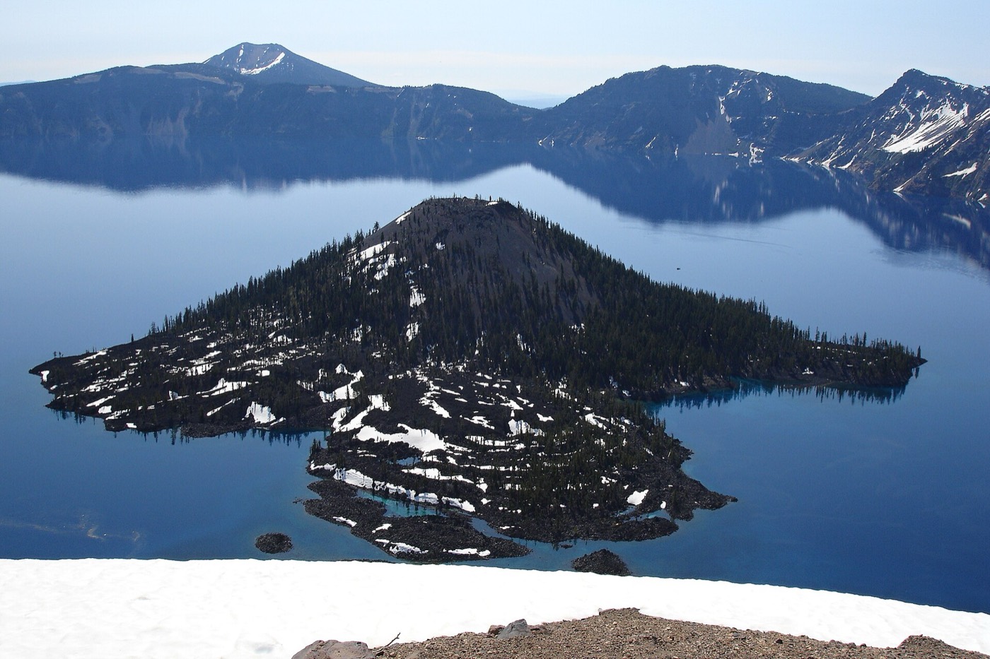 A wide modern view of Crater Lake from the rim, deep blue water ringed by cliffs with Wizard Island at the left, under an open sky.