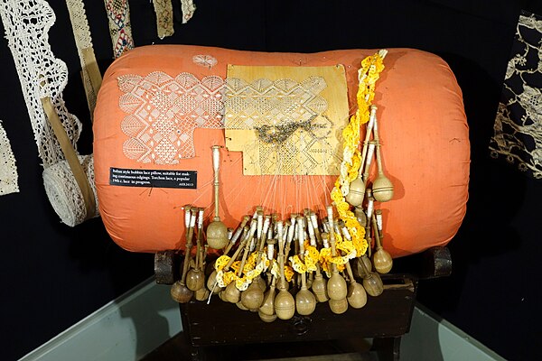 Bobbin lace in progress on an Italian-style pillow, with dozens of wooden bobbins and pins holding the emerging torchon pattern.