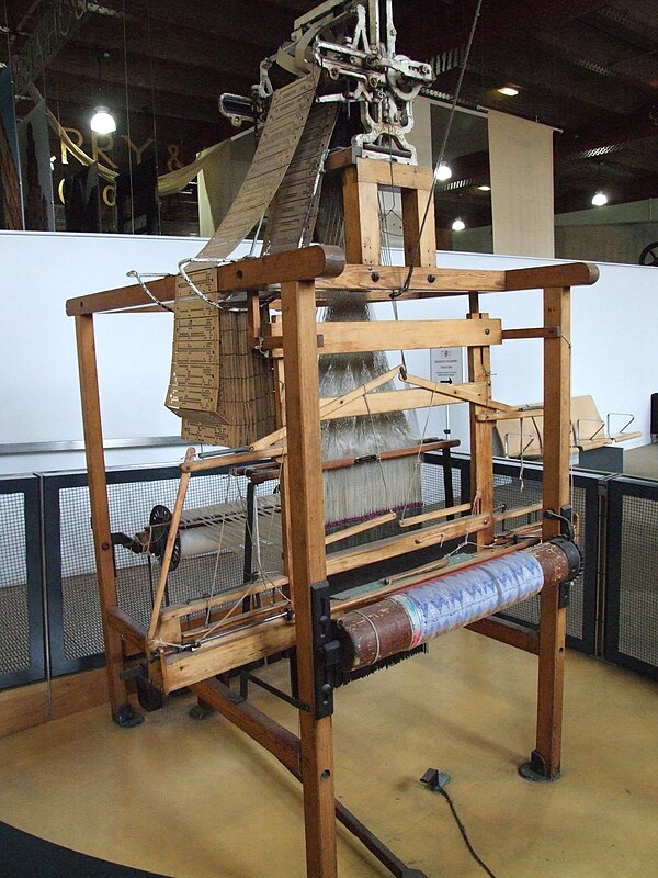 A wooden Jacquard loom with its chain of punched pasteboard cards visible at the top of the mechanism.