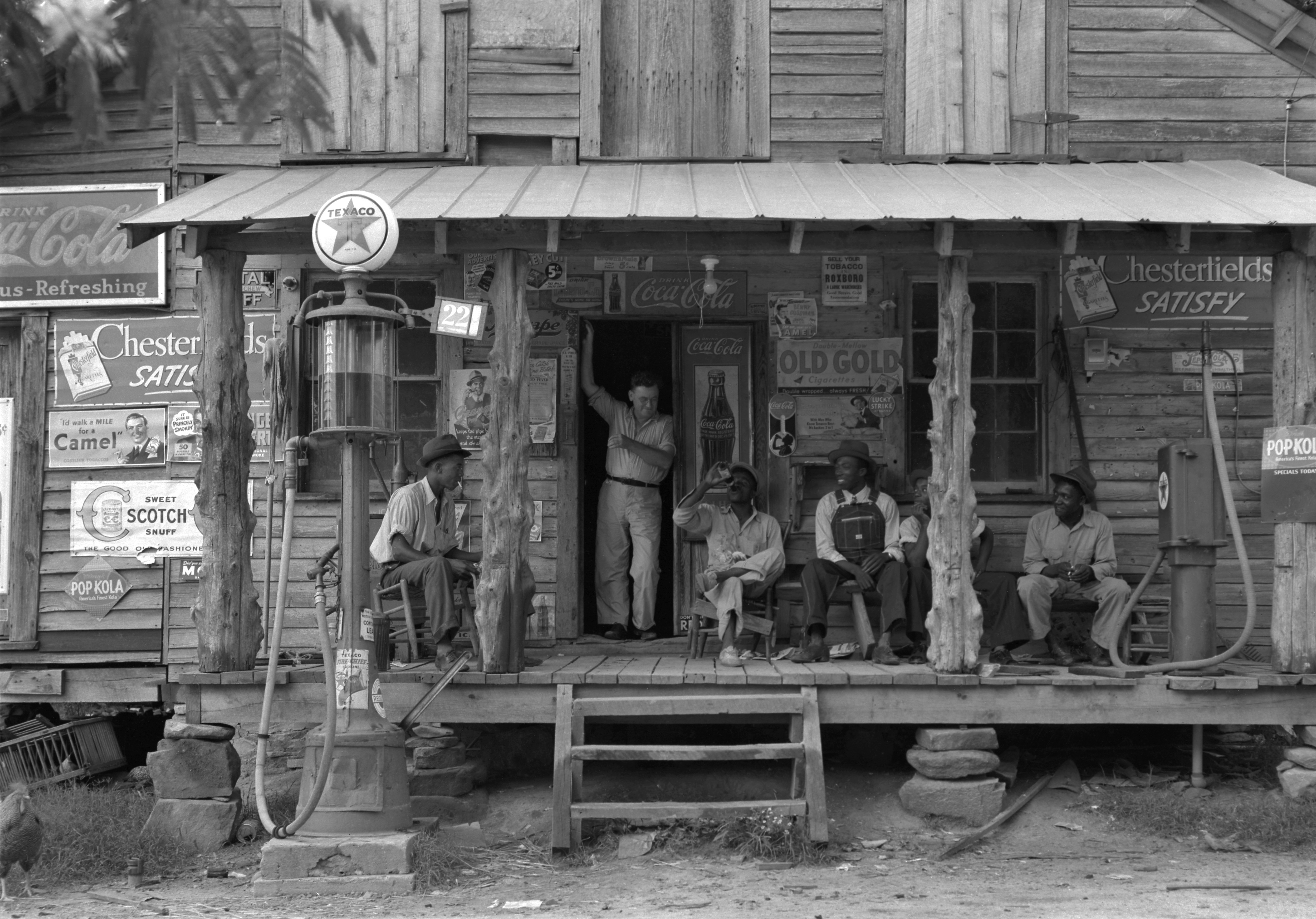 Five men sit on the porch of a rural country store on a dirt road, a kerosene pump on the right and a gasoline pump on the left.