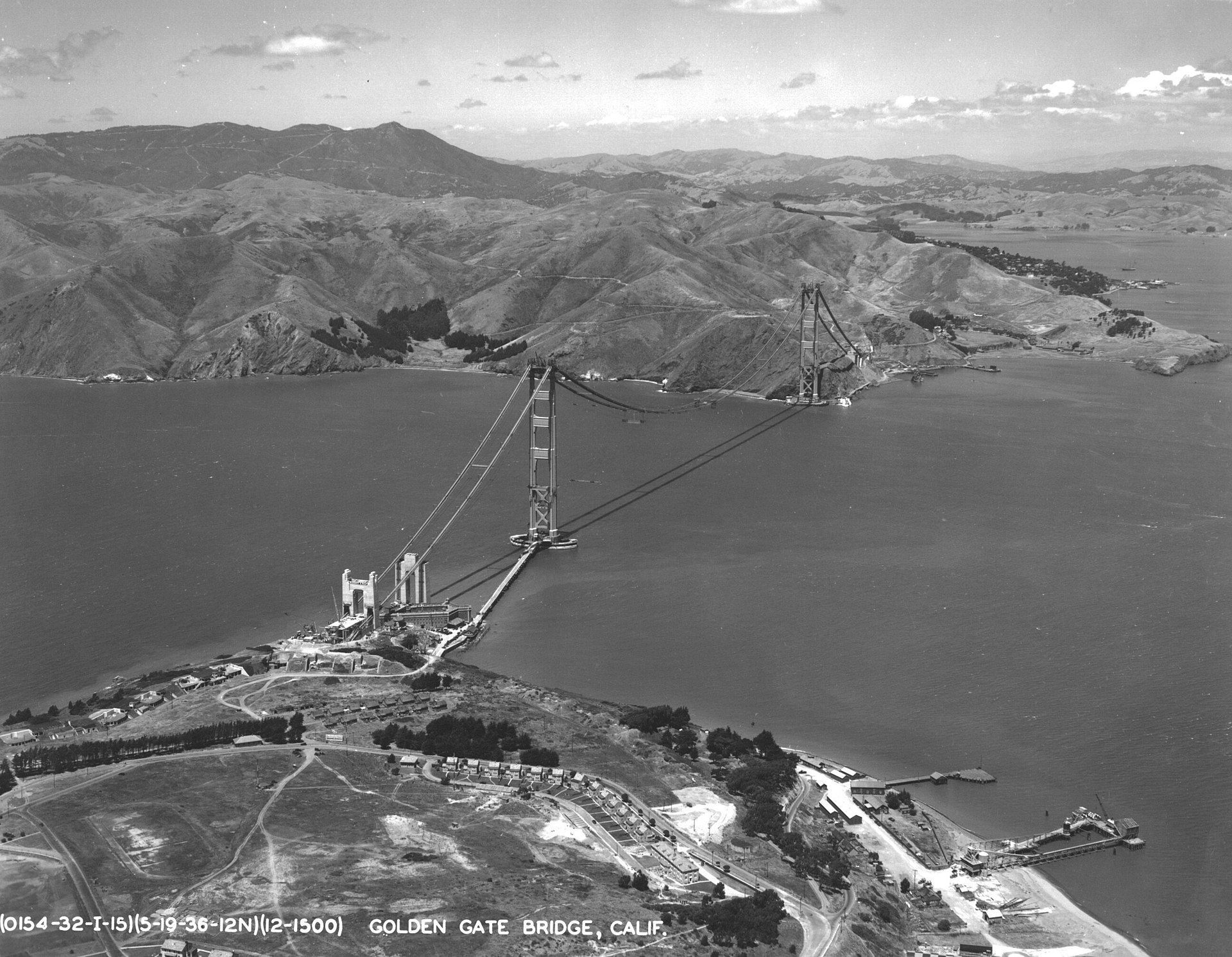 The south tower of the Golden Gate Bridge rising from the water, half-built, with partial catwalks visible, photographed from the air.