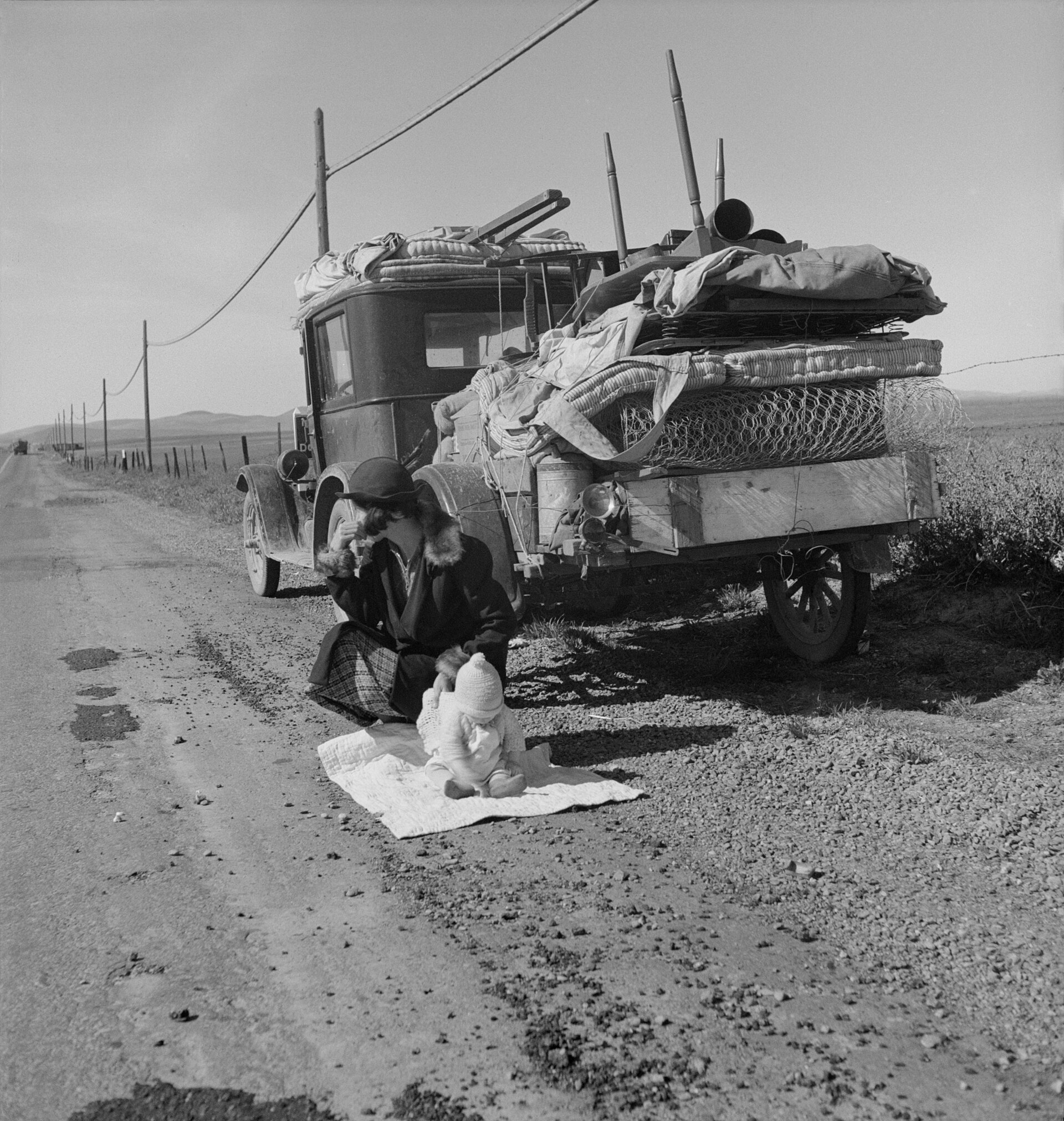 A Depression-era family of five clustered next to a broken-down car on the side of a California road, mother holding an infant.