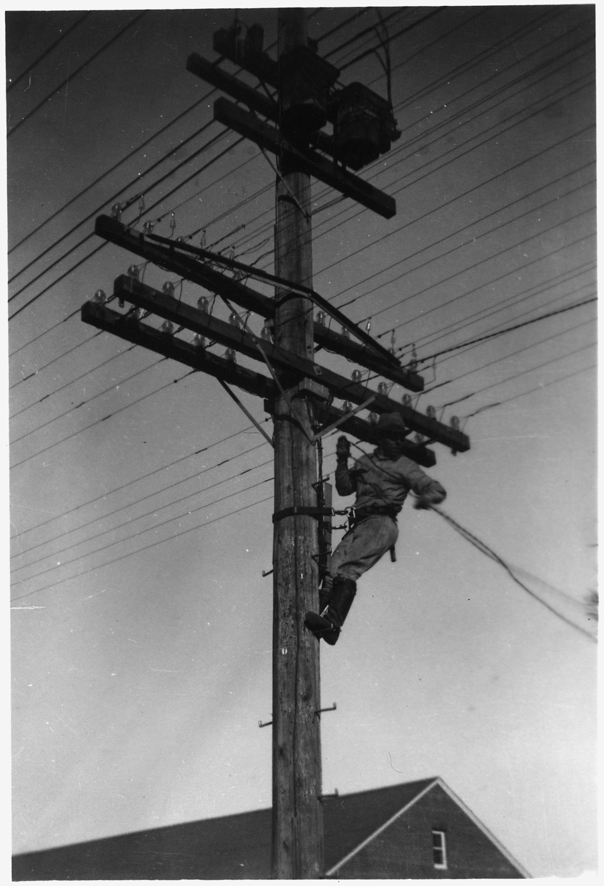 A lineman in work clothes climbs a wooden telephone pole against a plain sky, leather belt and climbing spurs visible.
