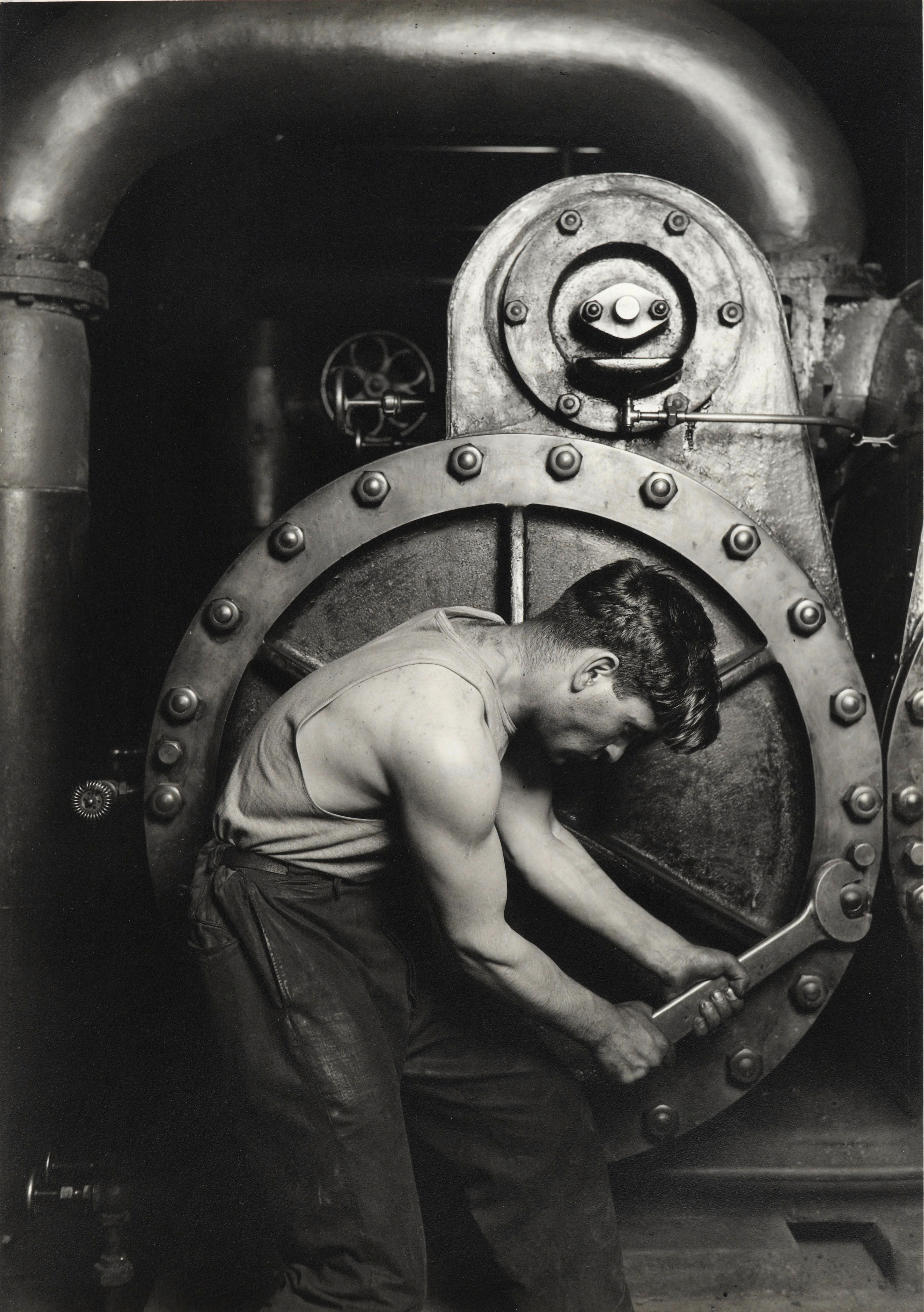 A muscular mechanic in dirty overalls bends around a massive steam-pump valve, wrench in hand.