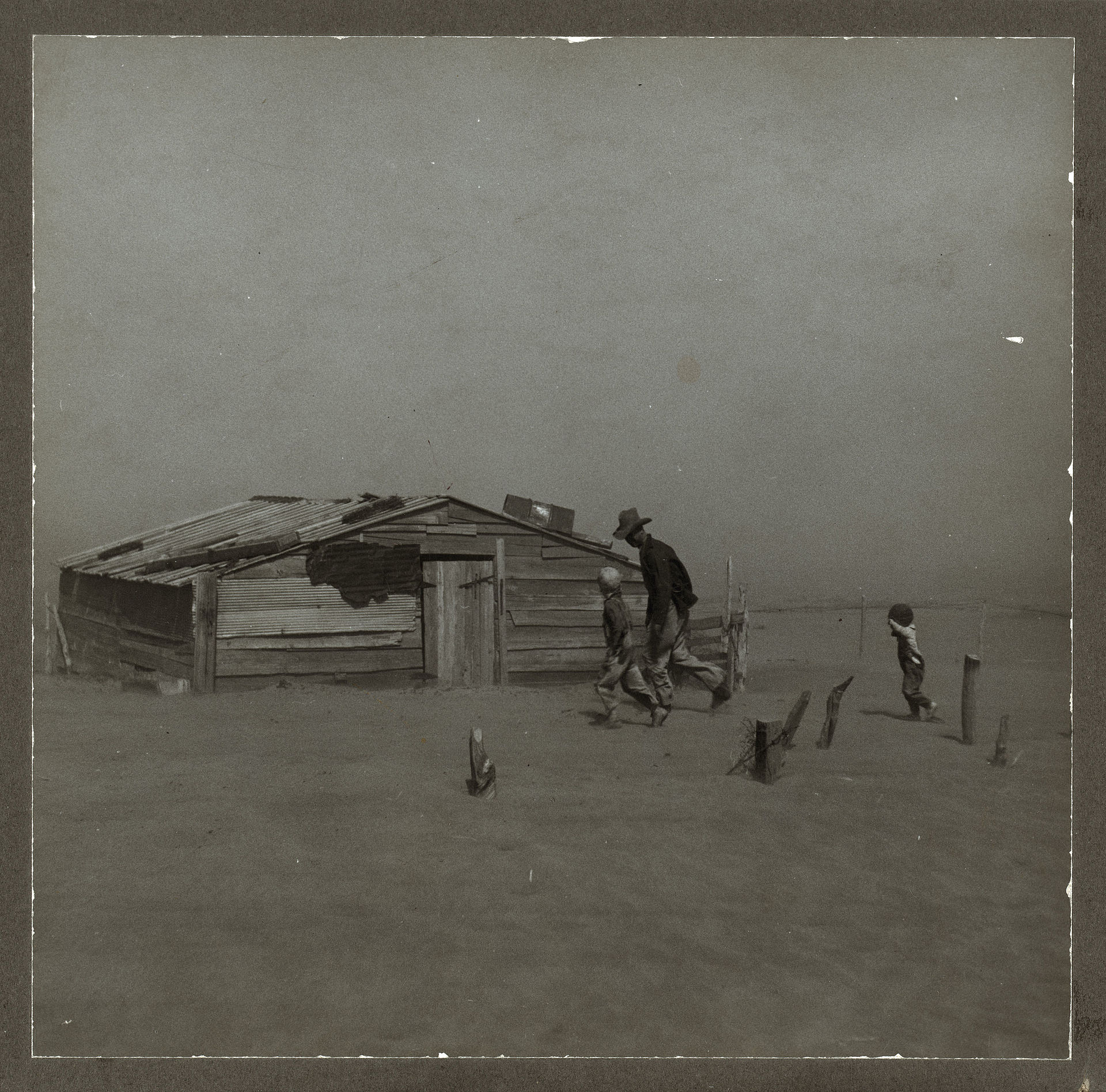A farmer and his two young sons walking away from a wooden shed, hunched against a rolling dust storm on the Oklahoma plains, 1936.