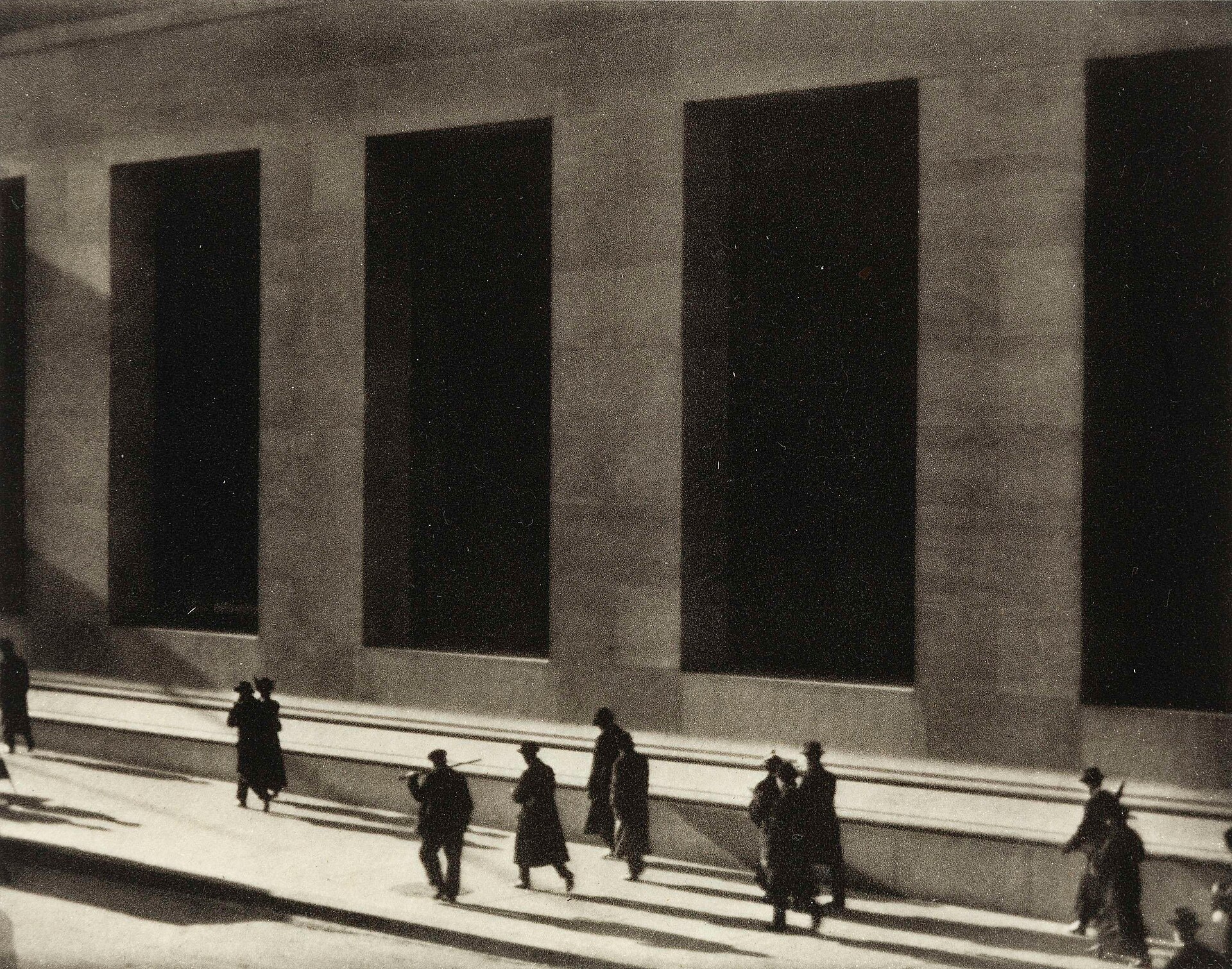 Small bowler-hatted figures walking past the shadowed, massive stone columns of J.P. Morgan and Co. on Wall Street, photographed in 1915.