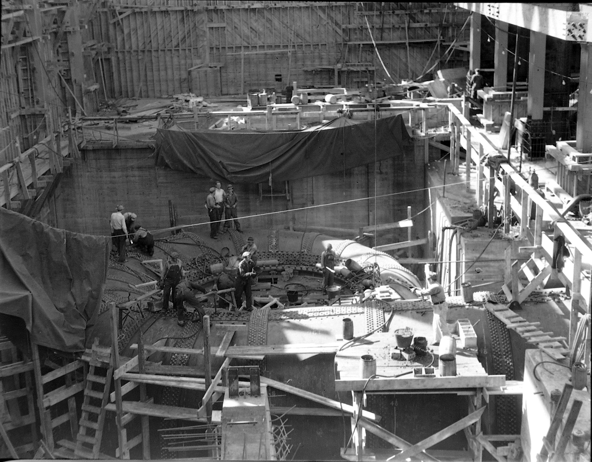 Construction workers stand inside a large concrete powerhouse under construction, with rebar and formwork rising around them.