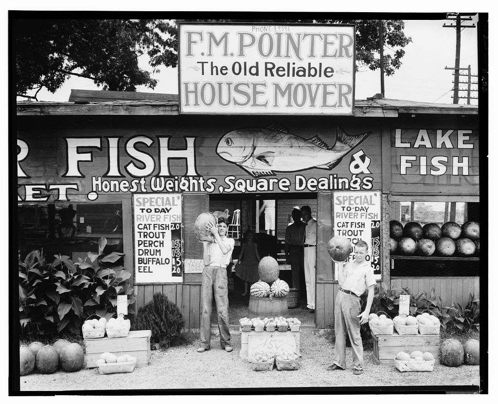 Two men holding watermelons in front of a wooden roadside fish-and-produce stand near Birmingham, Alabama, painted with hand-lettered signs for fish prices, photographed by Walker Evans in 1936.