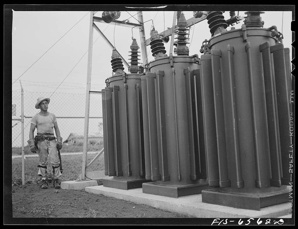 A Rural Electrification Administration lineman in coveralls and a hard hat examining the bushings of pole-mounted transformers at a small substation in Hayti, Missouri, 1942.
